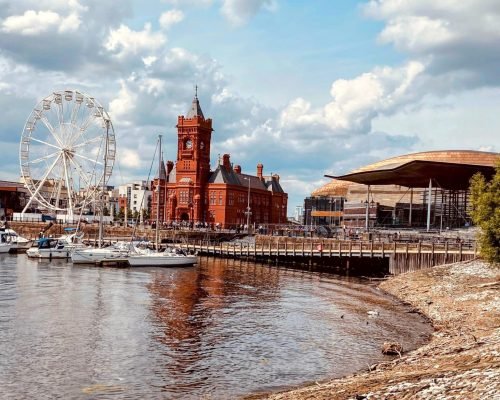 A vibrant view of Cardiff Bay’s Mermaid Quay, with sunlit waterfront terraces lined with restaurants and cafés, people strolling along the boardwalk, and boats gently bobbing in the water. The scene captures the lively atmosphere, modern architecture with maritime-inspired features, and the sparkling reflections of the bay under a clear blue sky.